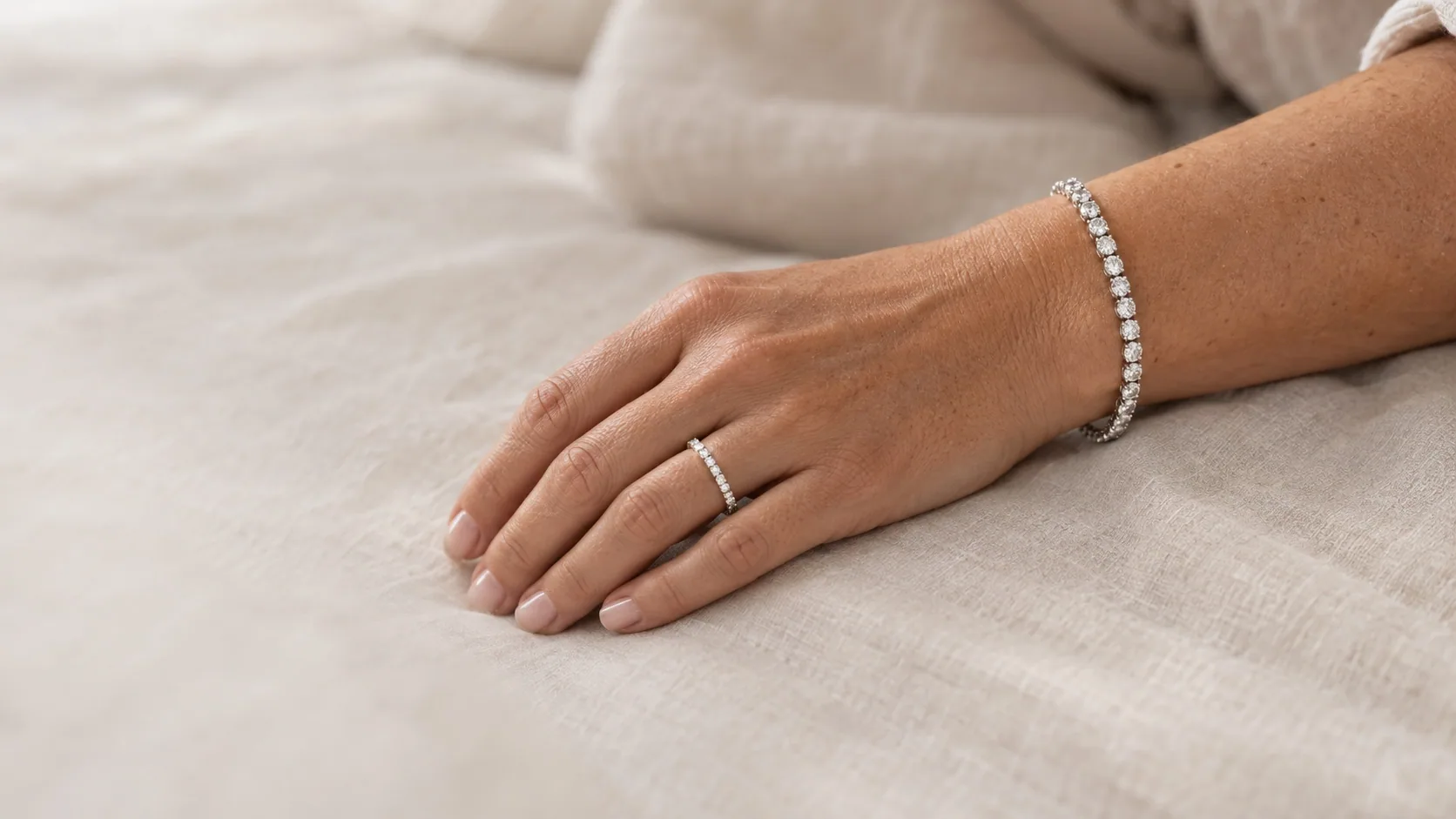 A diamond tennis bracelet and a diamond band worn on a hand resting on linen
