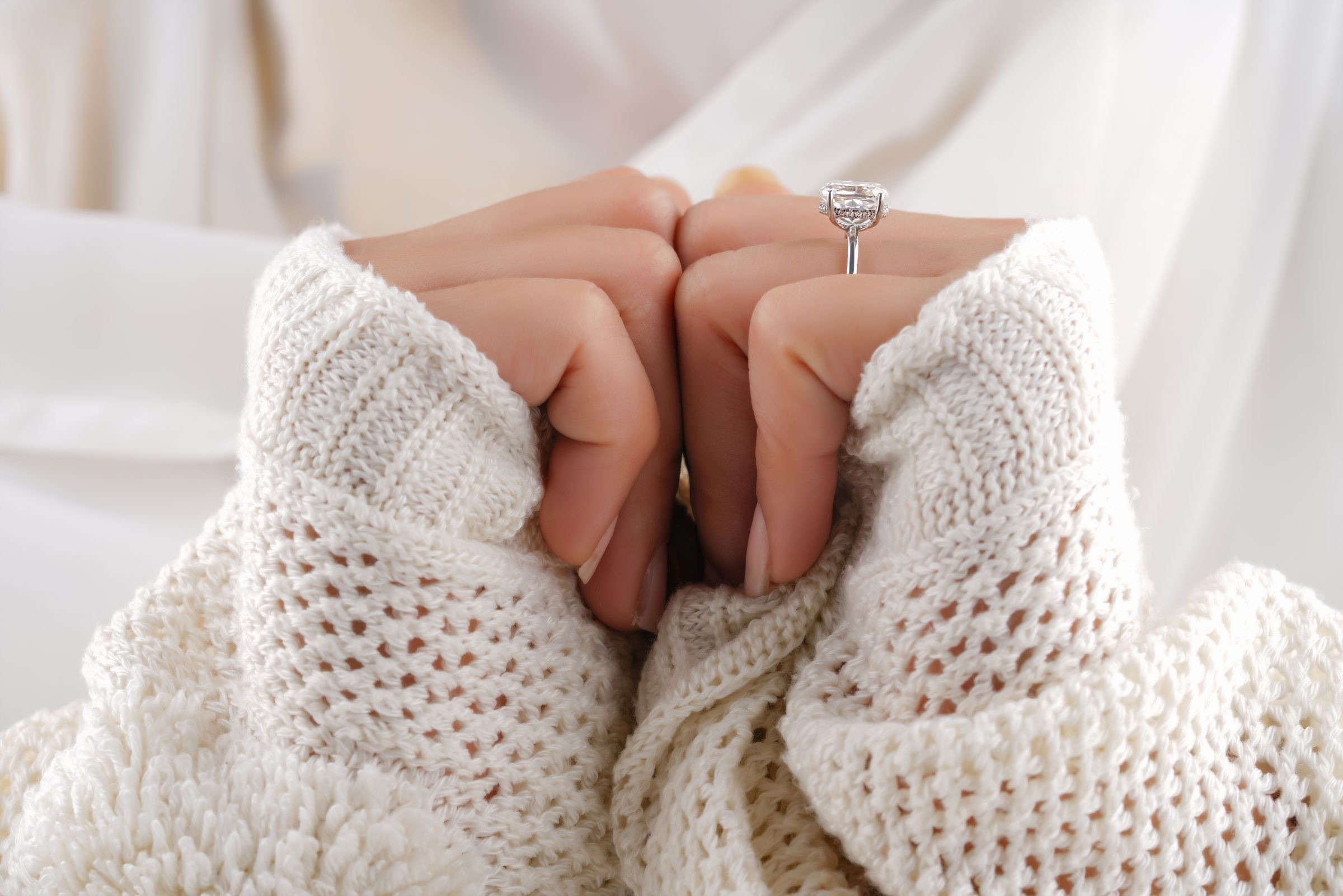 Two hands in cream knit sleeves, a diamond ring seen in profile